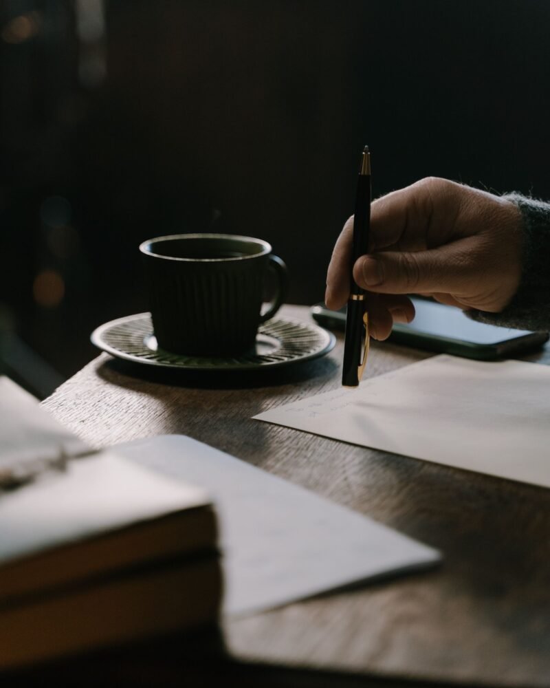 A hand holding a pen above a handwritten letter beside a coffee cup on a desk.