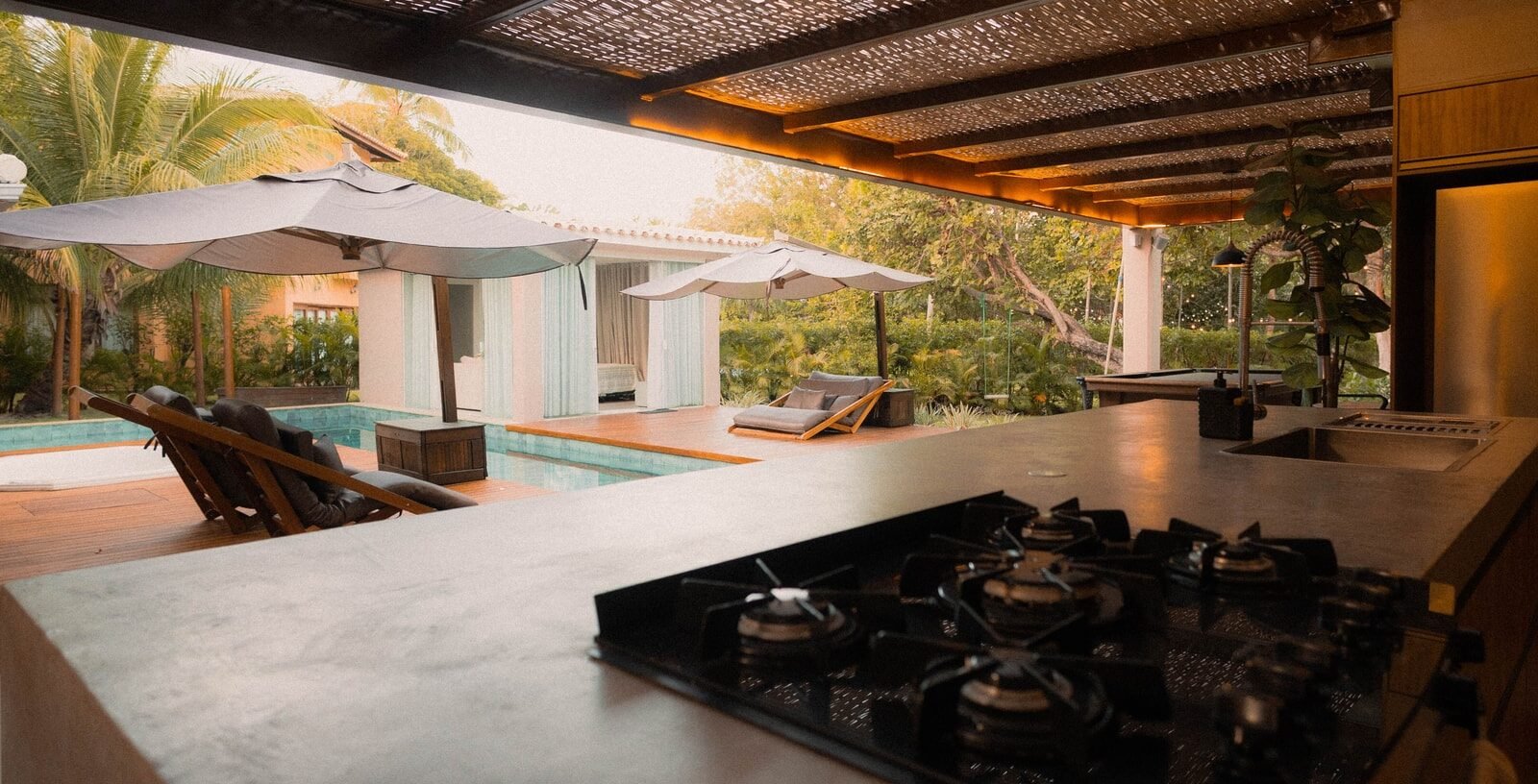 An outdoor kitchen under a woven pergola, looking out to a pool with umbrellas
