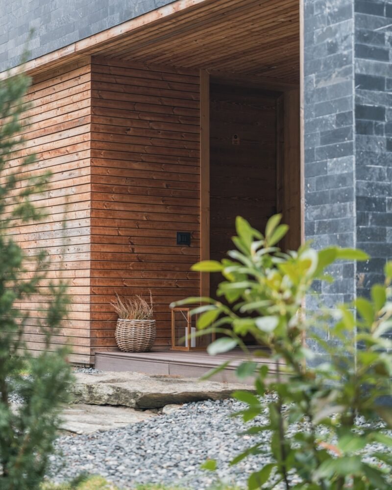 A covered entrance clad in timber and dark stone, framed by green plants