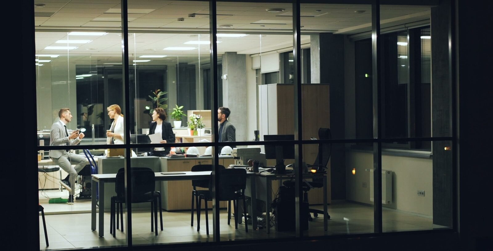 A lit office at night seen through tall windows, colleagues meeting inside