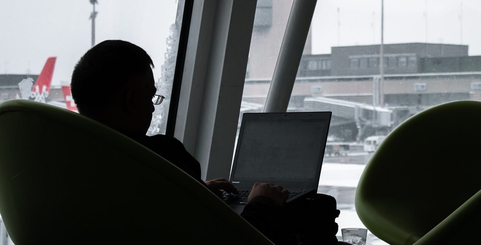 A man in silhouette working on a laptop in an airport lounge beside tall windows