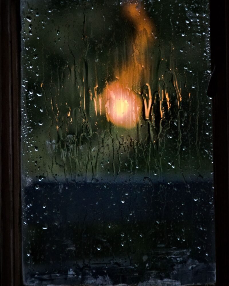Rain streaming down a window at night, a warm light blurred behind the glass