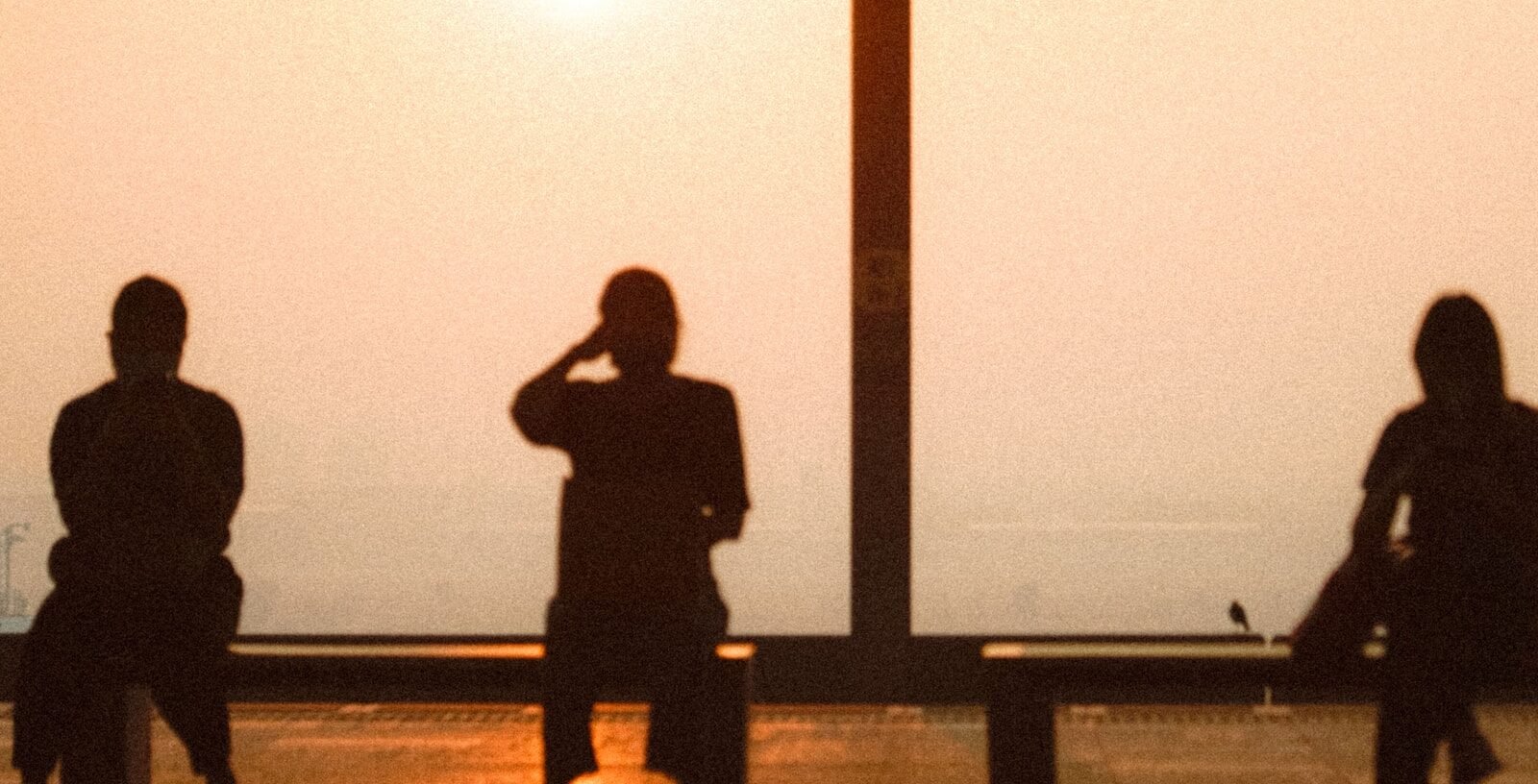 Three people silhouetted on a waterfront bench against a hazy orange sunset sky