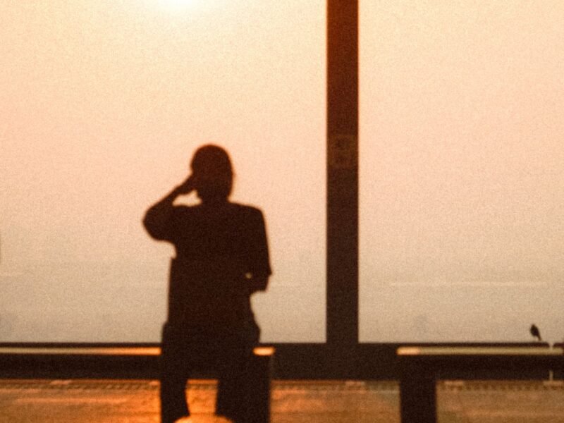 Three people silhouetted on a waterfront bench against a hazy orange sunset sky