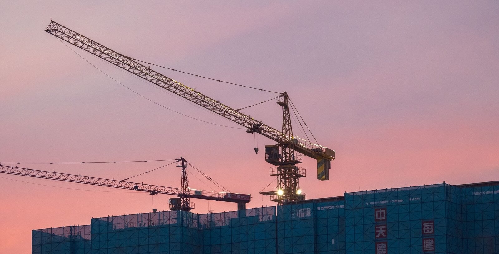 Tower cranes silhouetted against a pink dusk sky above a residential building wrapped in blue scaffolding netting
