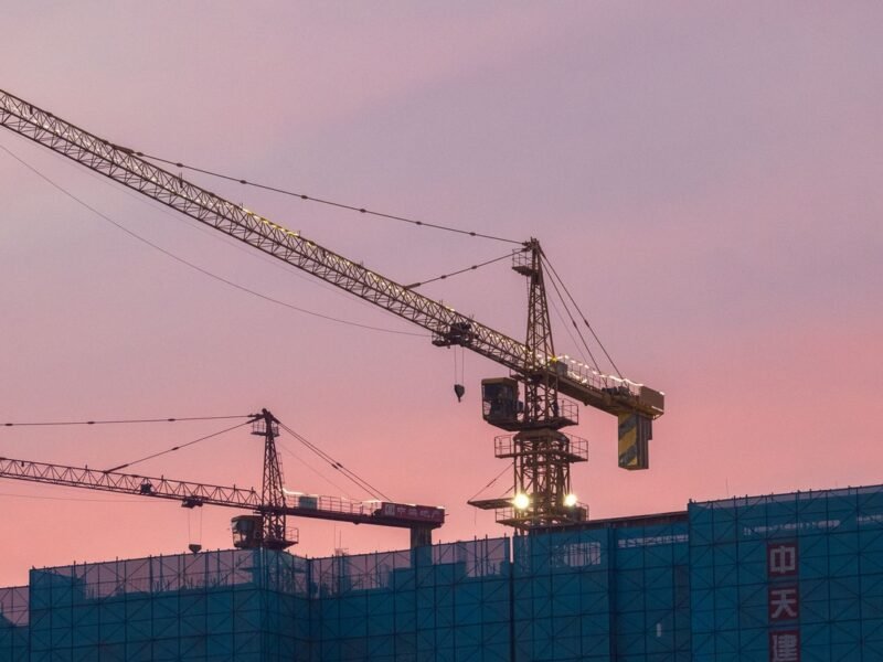 Tower cranes silhouetted against a pink dusk sky above a residential building wrapped in blue scaffolding netting